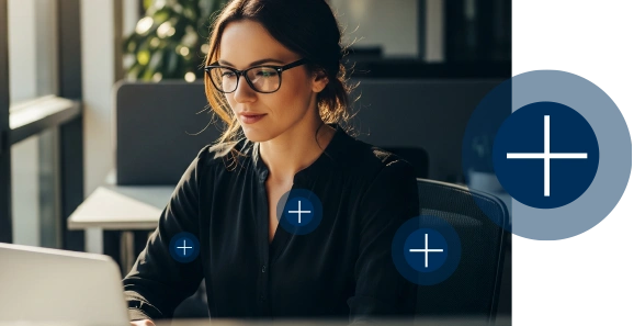 Professional woman wearing glasses working on a laptop at a desk in an office, with blue plus symbols floating around to indicate add-on features or tools.